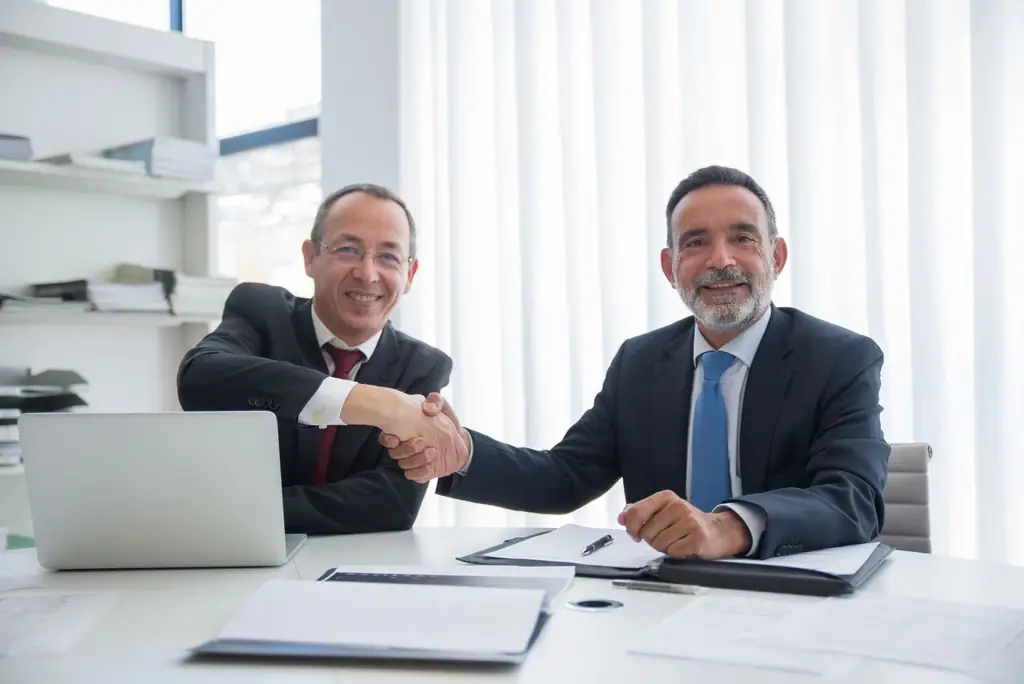 Two senior professionals shaking hands across a desk after an agreement